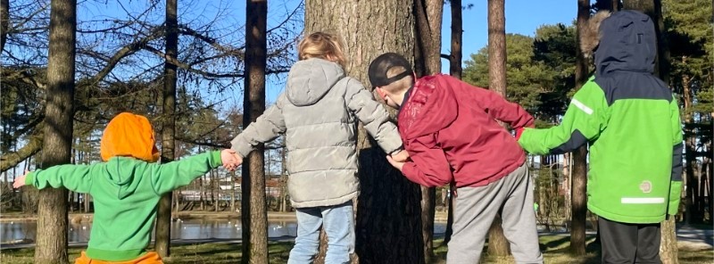 Tre born står ute på ein grøn eng, hand i hand, med ryggen vendt mot kamera. Dei har på seg varme jakker og ser ut til å nyte ein frisk dag ute i naturen, omgjeve av opne marker og spredde tre. Foto.